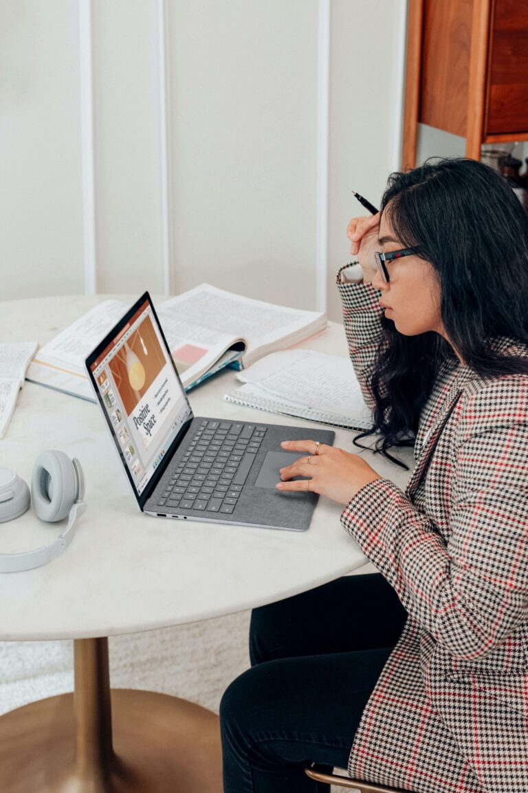 A woman sits at a round table using a laptop, surrounded by open notebooks and wearing glasses and a checked blazer. Headphones rest on the table.
