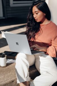 a young woman sits on a bench outside while using her laptop.