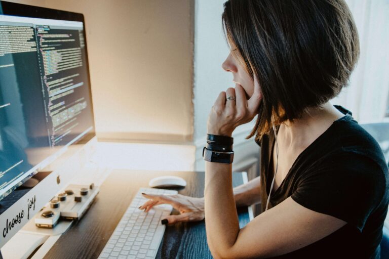 A person with shoulder-length hair sits at a desk, focusing on WordPress security as they look at code on a large computer monitor and type on a keyboard.