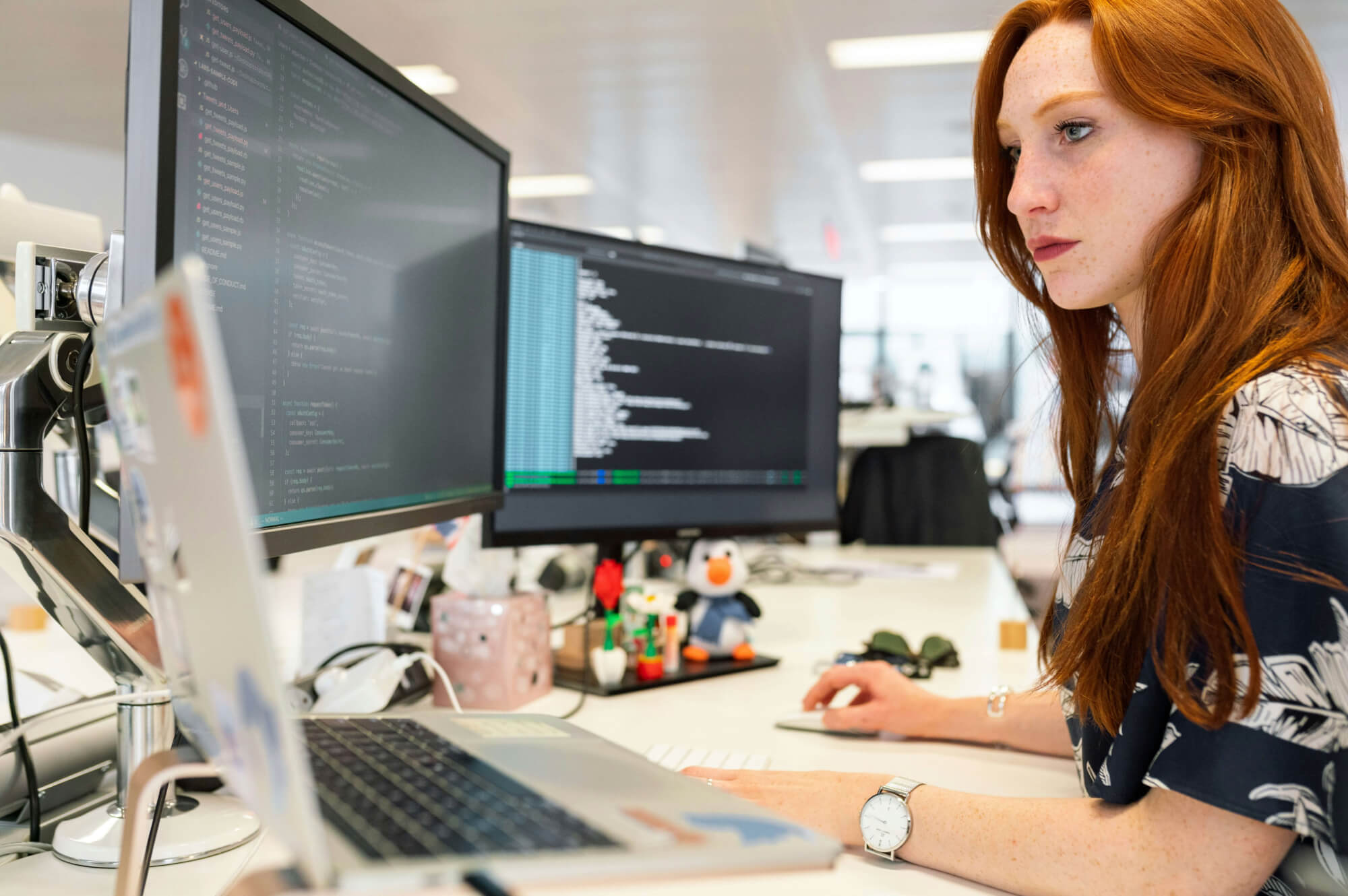 A woman with long red hair works at a desk with two computer monitors displaying code for building themes for WordPress, a laptop, and office supplies. She appears focused while typing in a bright, modern office environment.