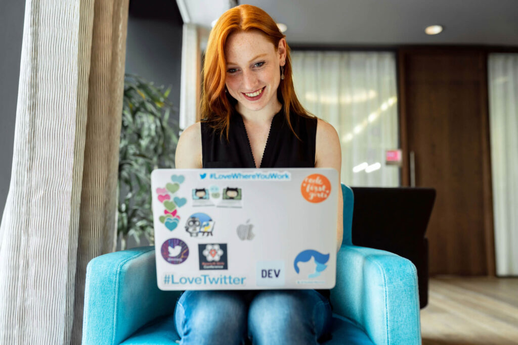 A smiling woman with red hair sits in a blue chair, working on a sticker-covered laptop. She appears focused and content in a modern, well-lit indoor space with plants and large windows.