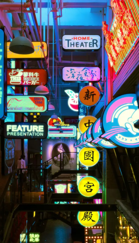 A narrow alleyway illuminated by colorful neon signs in various languages, including English and Chinese, advertising businesses like a home theater and feature presentation.