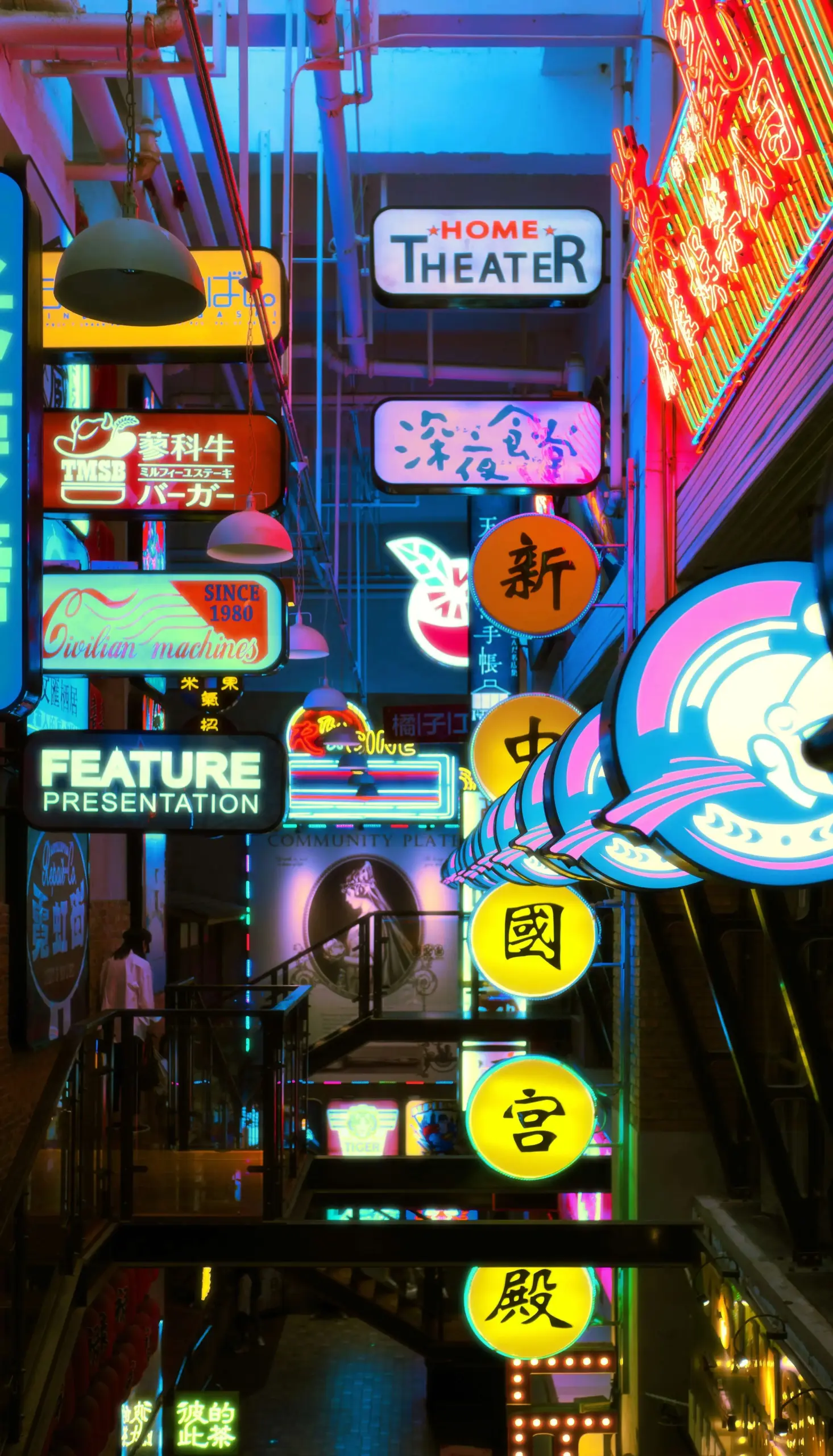 A narrow alleyway illuminated by colorful neon signs in various languages, including English and Chinese, advertising businesses like a home theater and feature presentation.