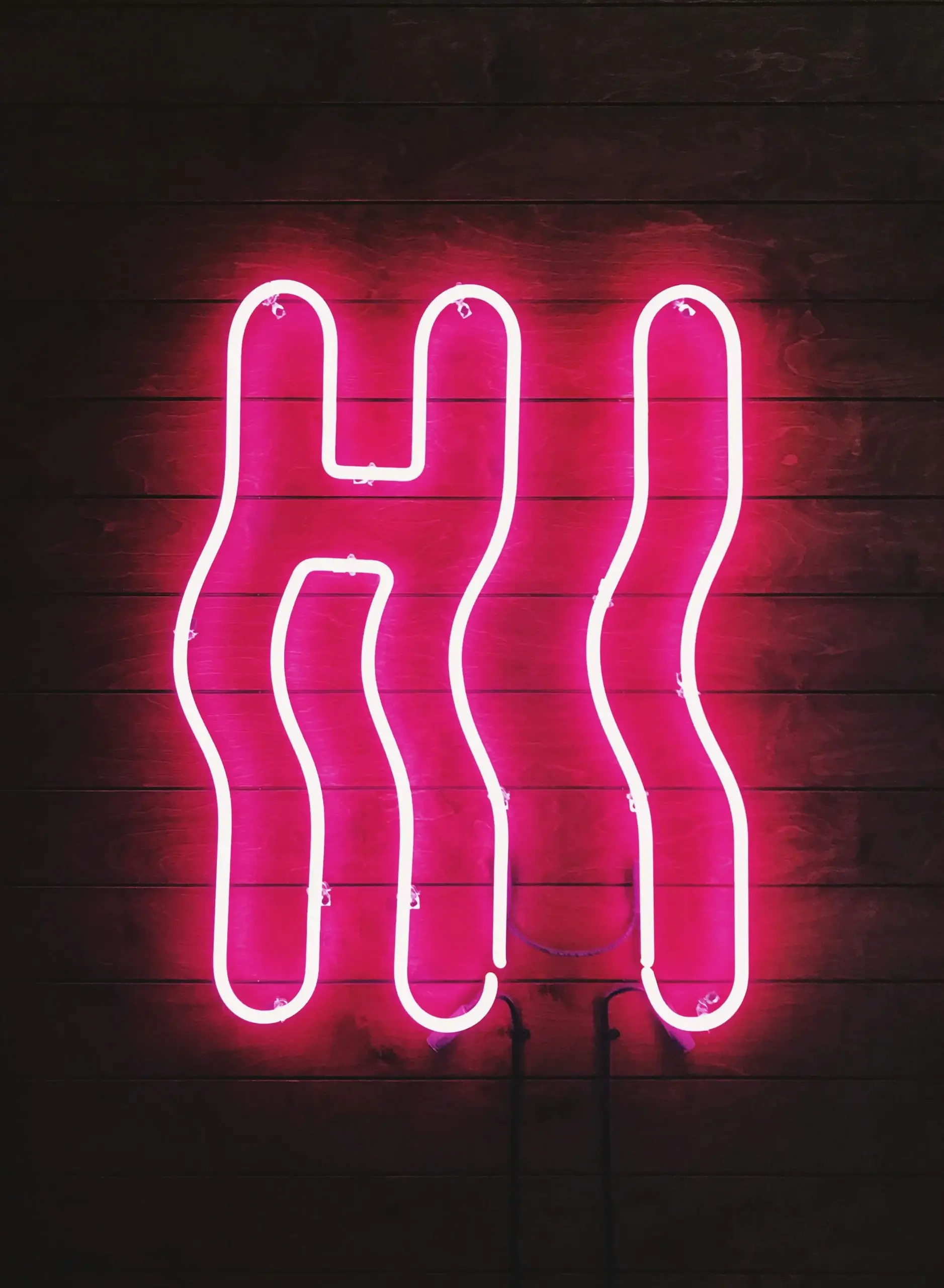 Pink neon sign shaped to spell “HI” on a dark wooden background.