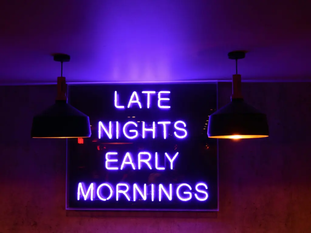 A neon sign with purple light reads LATE NIGHTS EARLY MORNINGS, mounted on a wall with two black hanging lamps on either side.
